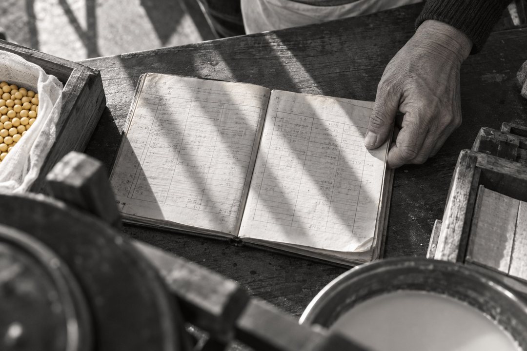 寄り。作業台の上に開いた古い大学ノート。鉛筆の細かい表が何十ページも続く(字は読めない)。ノートの端を押さえる、節の太い源治の指。